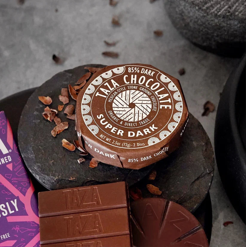A round tin of Taza Chocolate 85% Dark Super Dark chocolate sits on a dark stone slab, surrounded by chocolate pieces, cocoa nibs, and a purple package edge on a gray surface.