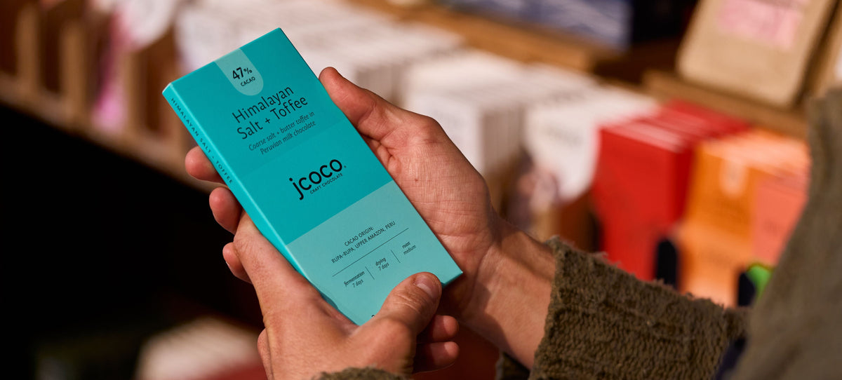 A person holding a turquoise blue Jcoco Himalayan Salt + Toffee chocolate bar in their hands, with a blurred store background.