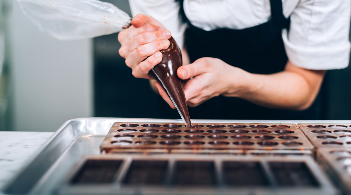A close-up of a person's hands holding a piping bag filled with dark chocolate and filling a mold on a baking tray.