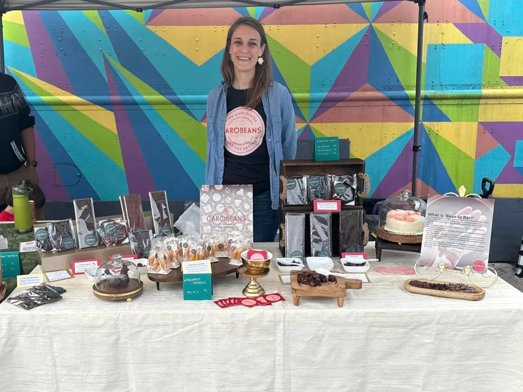 Owner of Carobeans Chocolate company in front of table with her products against a multicolor graphic background