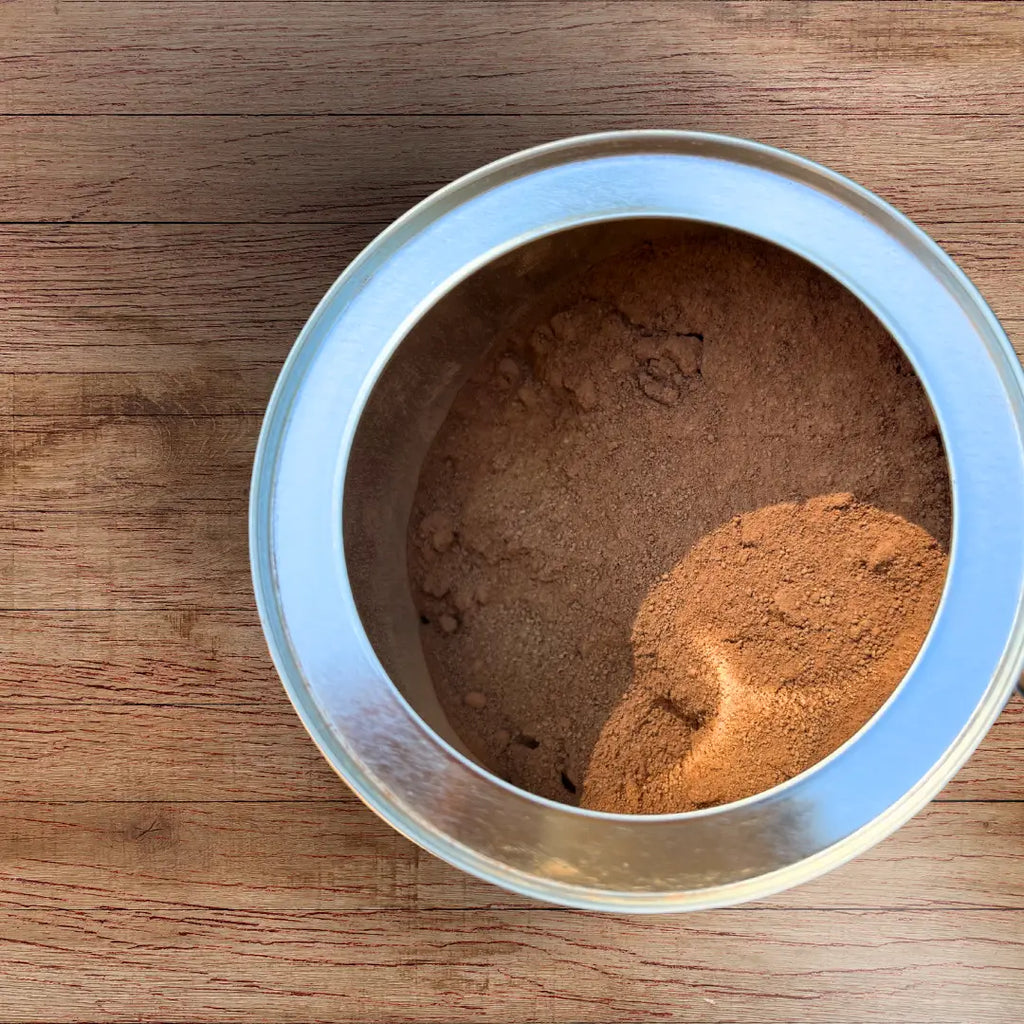 An open container of Ora Provisions Triple Fat Cacao Powder on a wood surface