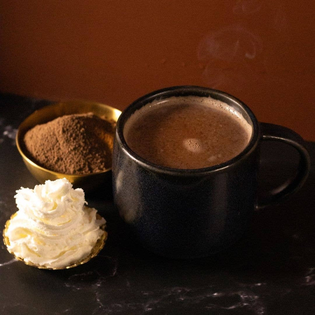 A black mug containing Cozy Dark Hot Chocolate on a marble table with cocoa powder and whipped cream