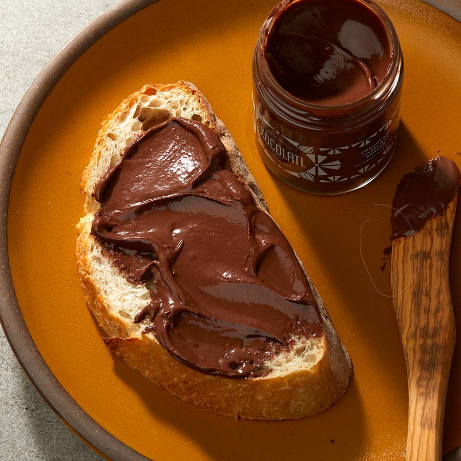 Chocolate Hazelnut Spread on a piece of break next to a jar of Chocolate Hazelnut Spread, all on an orange plate with a wooden spreading knife