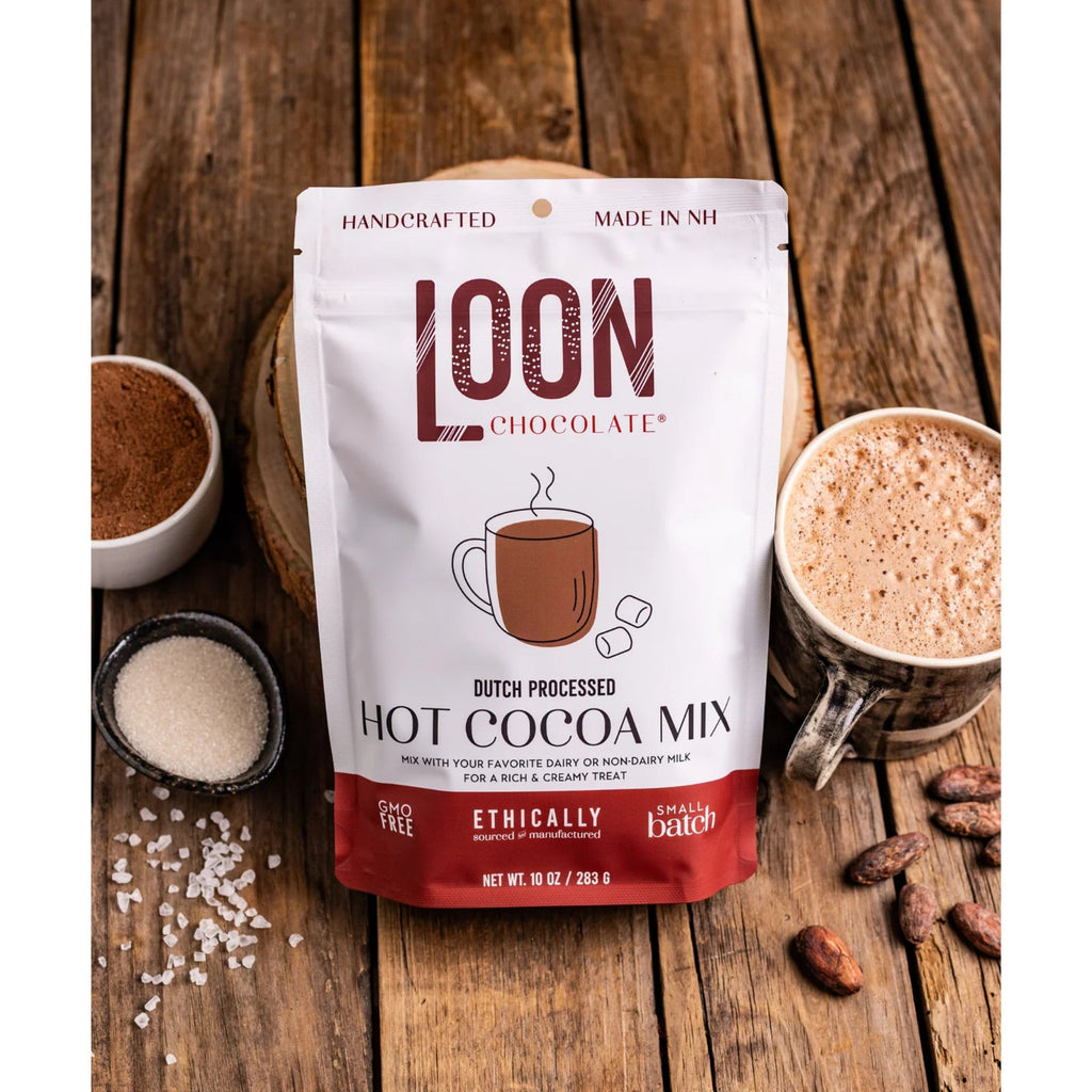 A bag of Hot Chocolate Mix from Loon Chocolate resting against cups of hot cocoa on a wooden table