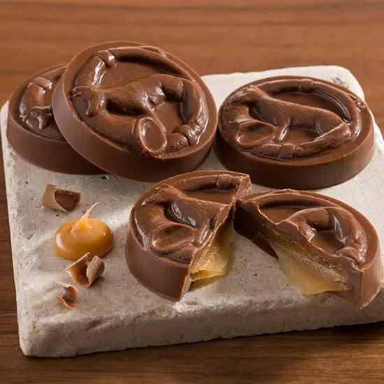 Four unwrapped Milk Chocolate Caramel Fox Trot chocolates on top of a cermic tile. One chocolate is cut in half and shows caramel spilling out. All four chocolates on a wood surface