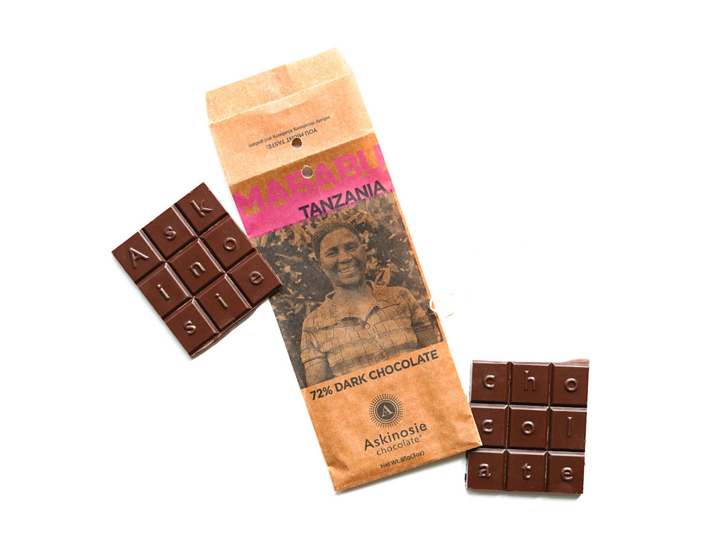 The front of a wrapped 72% Mababu, Tanzania Dark Chocolate Bar from Askinosie Chocolate, with the unwrapped bar broken into two parts beside the wrapping on either side, with a white background.