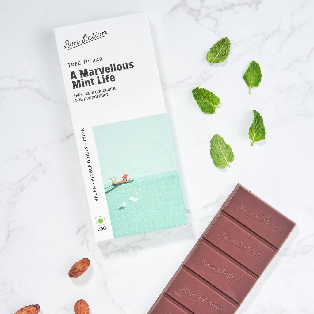 The front of a wrapped Marvelous Mint Life Bon Fiction Bar, adjacent to an unwrapped Marvelous Mint Life Bon FIction Bar, with mint leaves and cocoa beans on either side, against a marble background.
