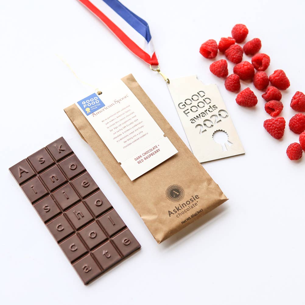 An unwrapped Dark Chocolate + Red Raspberry Bar, to the left of its wrapping, with raspberries and a "Godo Food Awards 2020" medal to the right of the wrapping, with a white background.