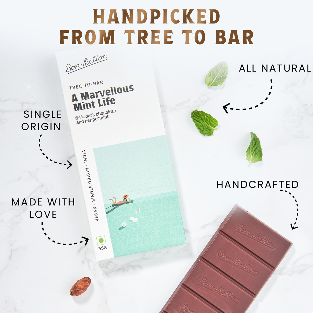 The front of a wrapped Marvelous Mint Life Bon Fiction Bar, adjacent to an unwrapped Marvelous Mint Life Bon FIction Bar, with mint leaves and cocoa beans on either side, against a marble background. There is an "Single-Origin" sign pointing to he middle of the wrapped bar, a "Made With Love" sign pointing to the bottom of the wrapped bar, an "All Natural" sign pointing to the mint leaves, and a "Handcrafted" sign pointing to the unwrapped bar. 