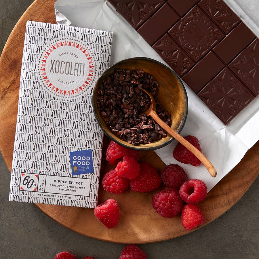 A wooden plate with a wrapped and unwrapped Ripple Effect: 60% Dark Chocolate with Raspberry & Nibs bar, a bowl of cocoa nibs, and raspberries
