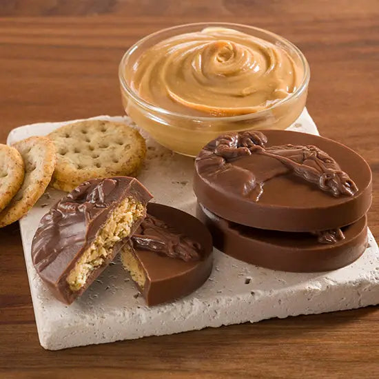 Unwrapped Milk Chocolate Peanut Butter Sea Biscuits  split in half to show peanut butter, bowl ofpeanut butter, crackers, all on a white tile on top of a wood background