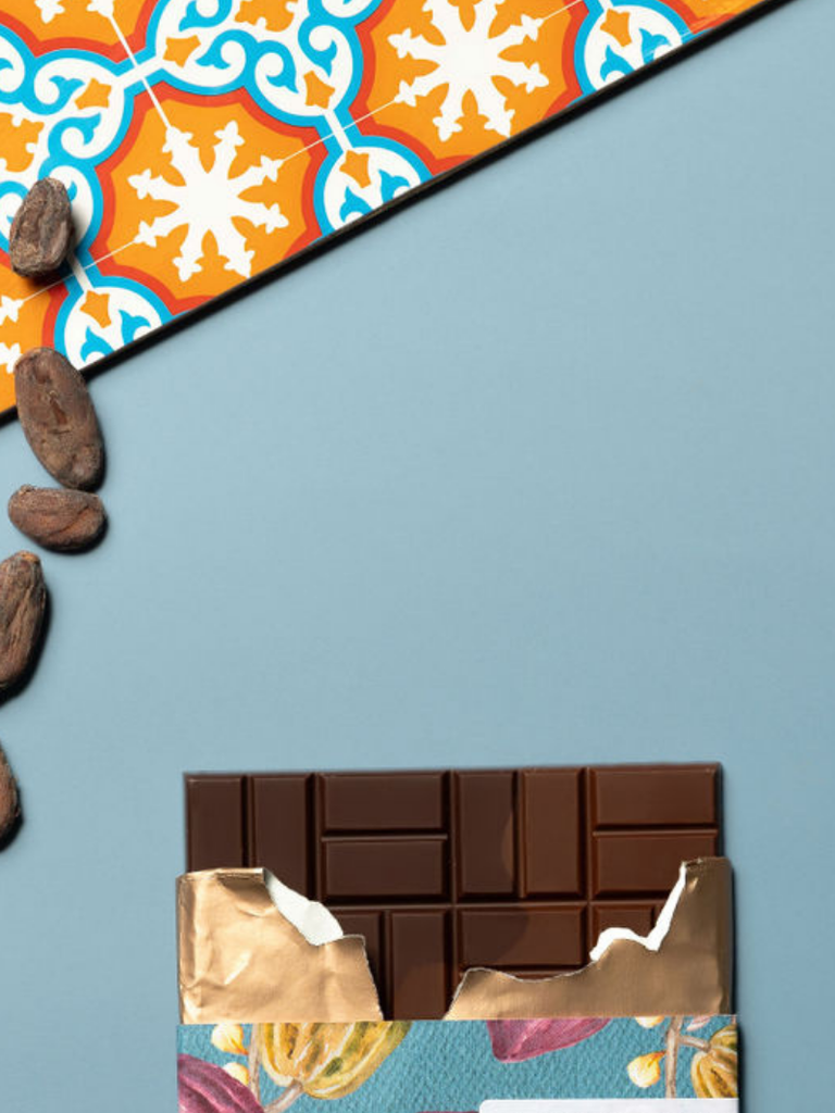 Partially opened Two Rivers 70% Dark Chocolate - Rio Negro e Rio Solimões bar on a blue table with a patterned cloth and cocoa beans