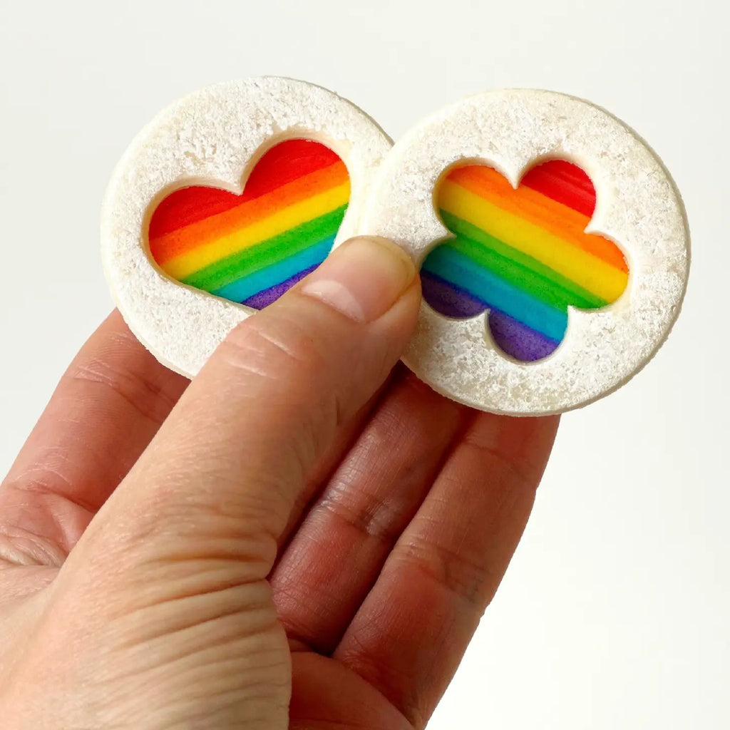 A hand holding up two unwrapped Marzipan Candy Pride Rainbow Linzer Cookies against a white background
