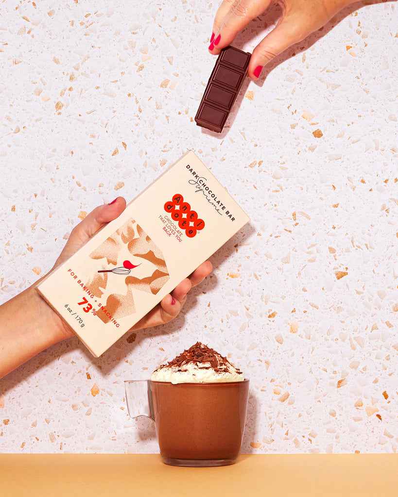 Two hands holding the Supreme: Bake + Snack 73% Dark Chocolate bar from Antidote chocolate above a cup of drinking chocolate against a patterned background