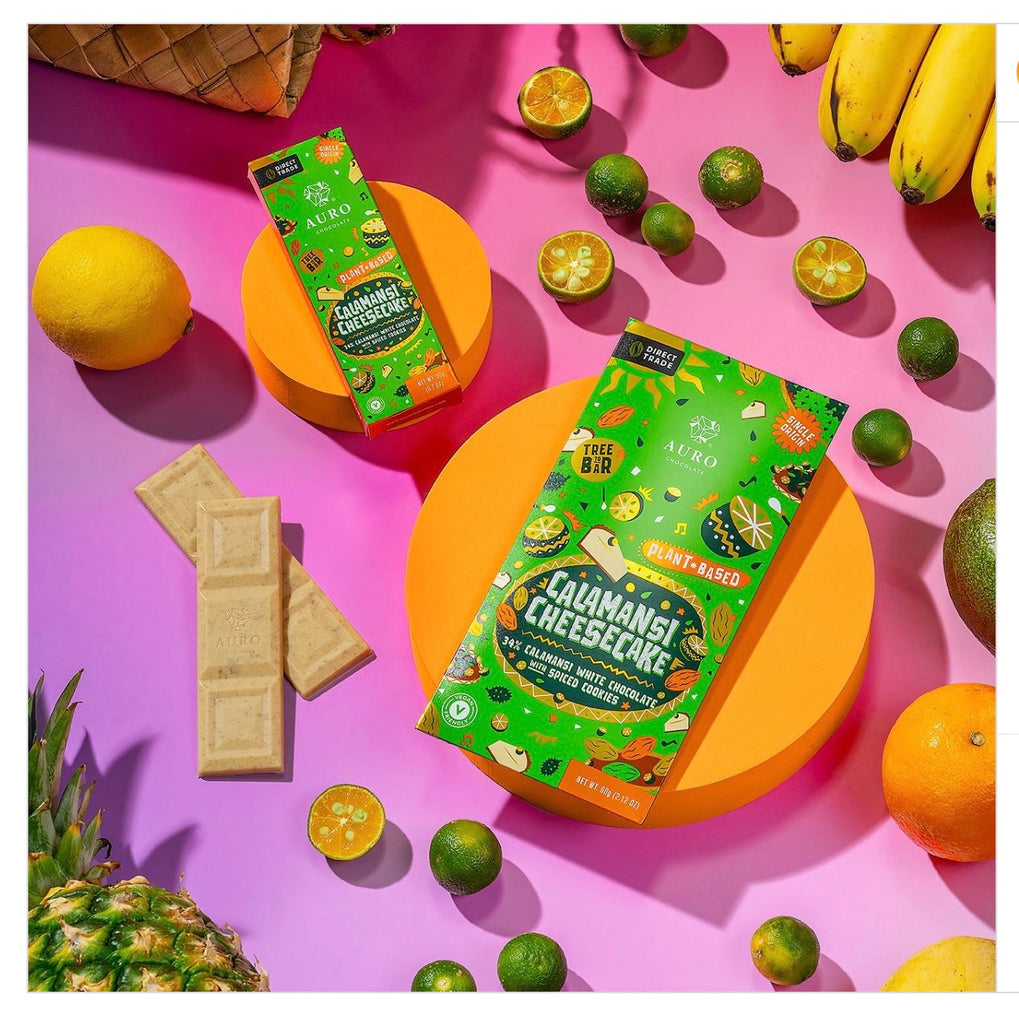 Two wrapped Auro White Chocolate Calamansi Cheesecake Bars and one unwrapped Auro White Chocolate Calamansi Cheesecake Bar with pineapple, lemons, and limes against a pink background