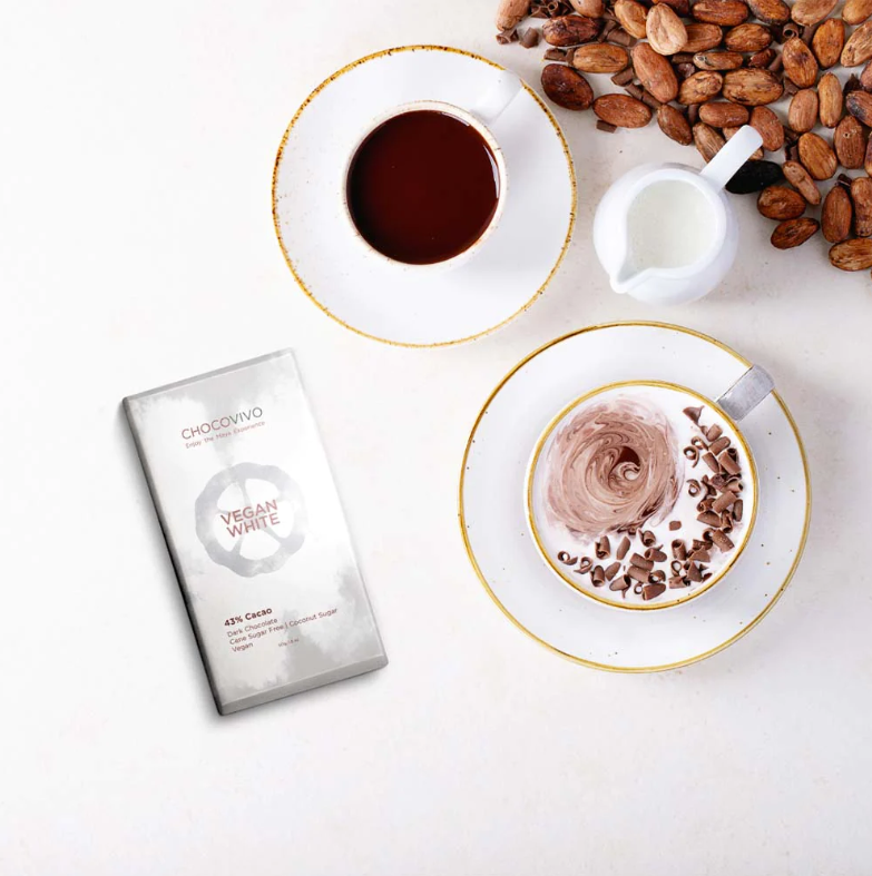 A ChocoVivo Vegan White 43% bar, two cups of drinking chocolate, a small pitcher of milk, and cocoa beans on a white table.