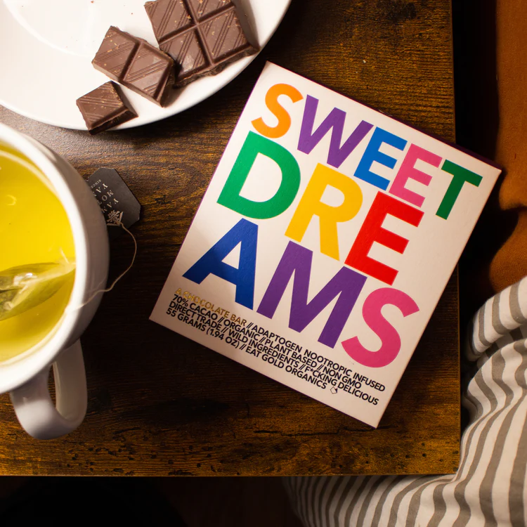 A wrapped Sweet Dreams chocolate bar on a table with a cup of tea and a plate of broken pieces of chocolate.