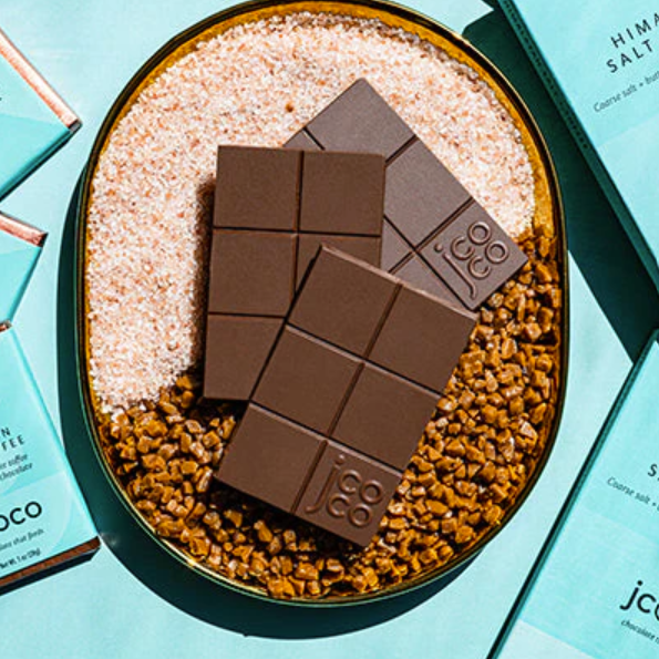 Three unwrapped Jcoco Himalayan Salt + Toffee bars on top of ingredients next to wrapped Himalayan Salt + Toffee bars on a light blue table