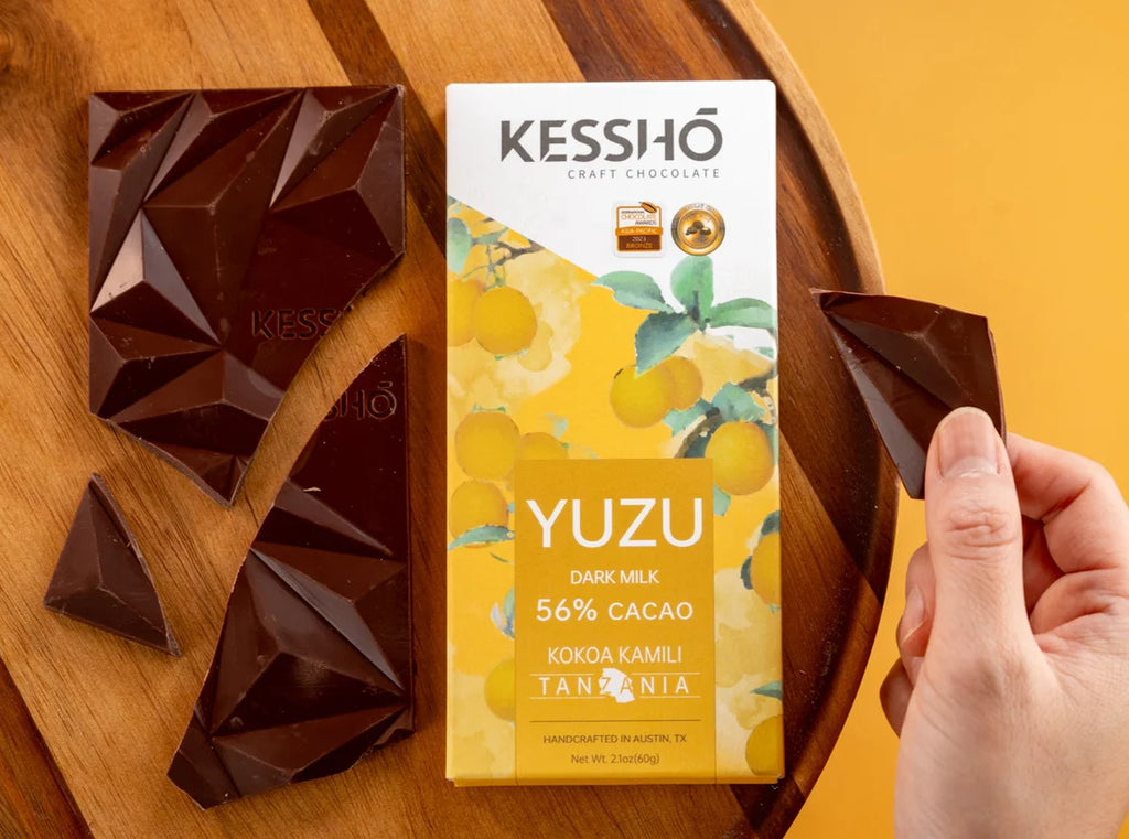 The front of a KESSHO wrapped Yuzu 56% Dark Milk back against a wooden background with broken pieces of a chocolate bar