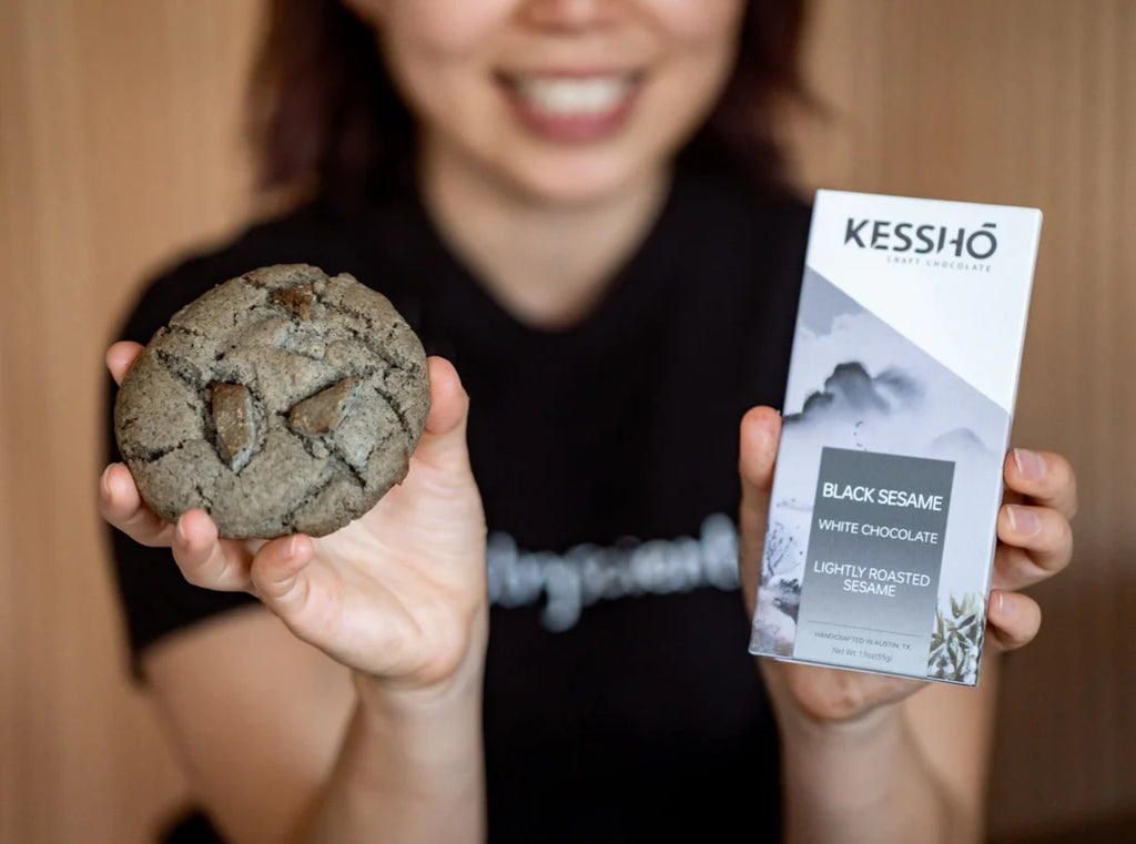 A smiling woman holding a wrapped Black Sesame White Chocolate bar in her left hand and a cookie in her right hand