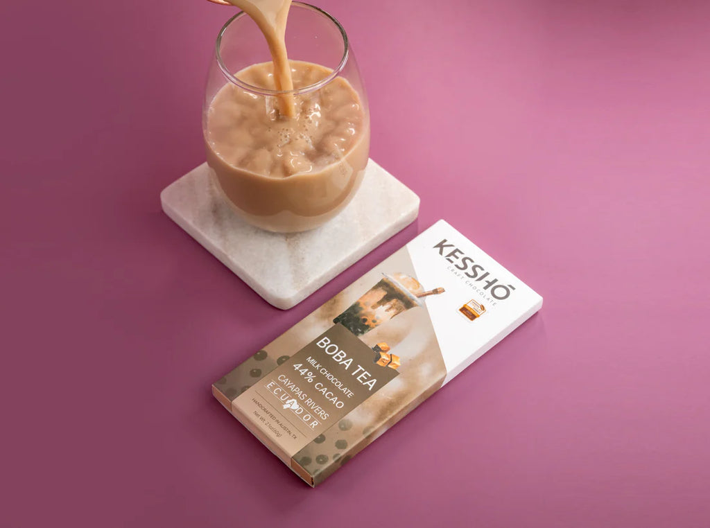 A wrapped Boba Tea 44% Milk Chocolate bar next to a beverage on a white coaster, on a pink table