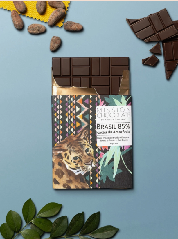 A partially opened 85% Cacao - Brazil bar on a blue table with unwrapped chocolate, cocoa beans, and leaves
