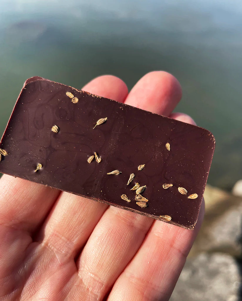 Hand holding a piece of an unwrapped Queen T: Anis + Tomato bar with the ocean in the background