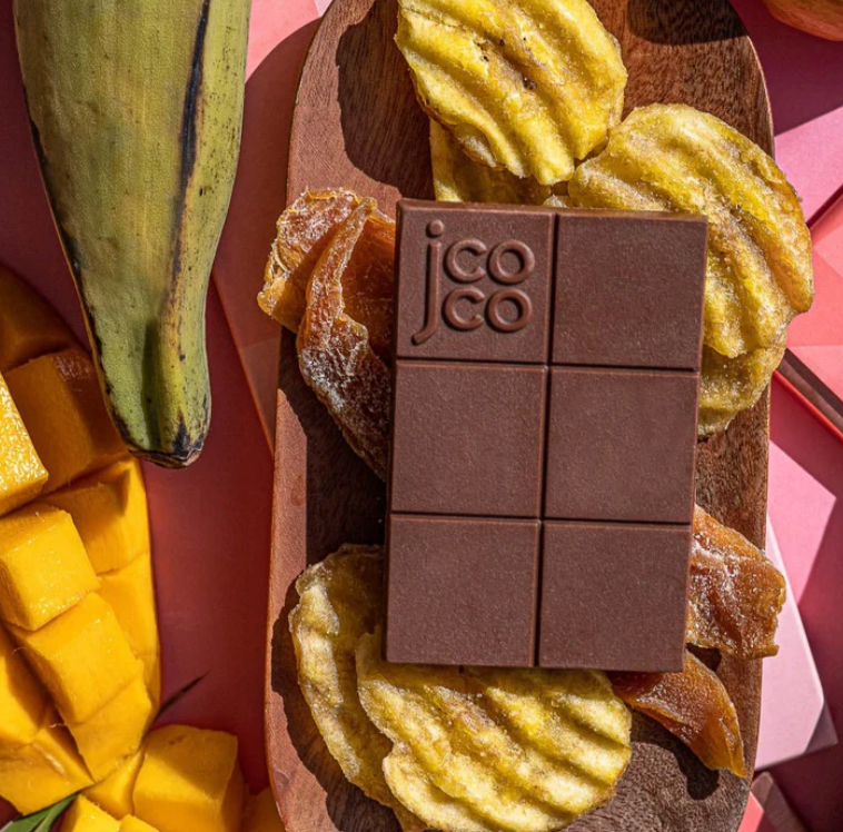 Unwrapped Mango Plantain Milk Chocolate 47% Mini on a wooden plate with plantain chips and dried mangoes.
