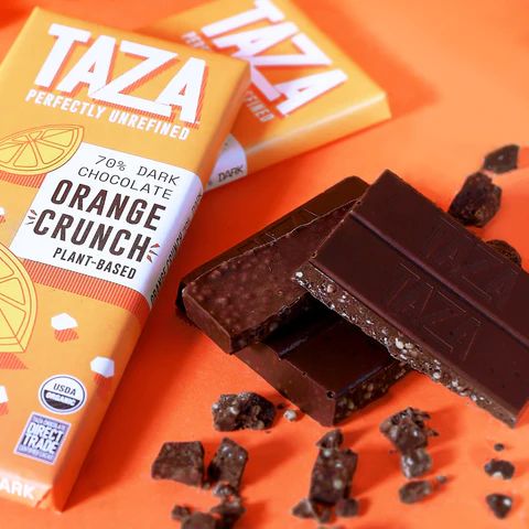 Two Orange Crunch 70% bars on an orange table with unwrapped pieces of Taza chocolate