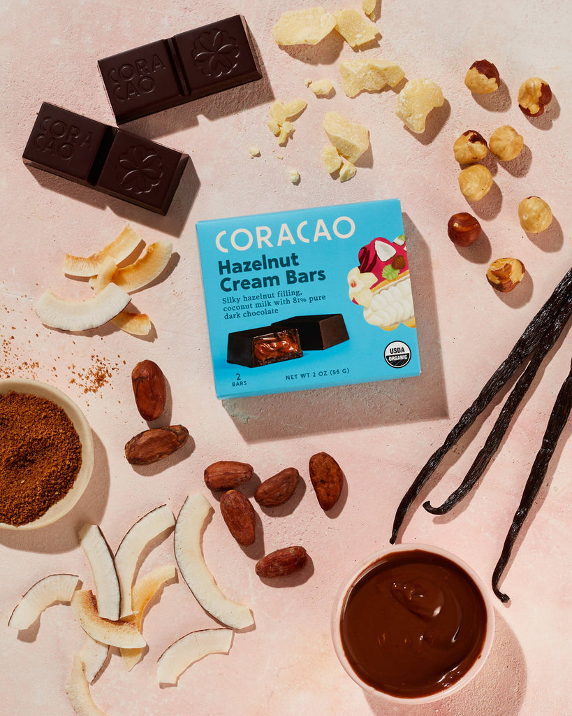 Vegan Hazelnut Cream Bar 2-Pack box with various unwrapped ingredients and pieces of chocolate on a light pink table