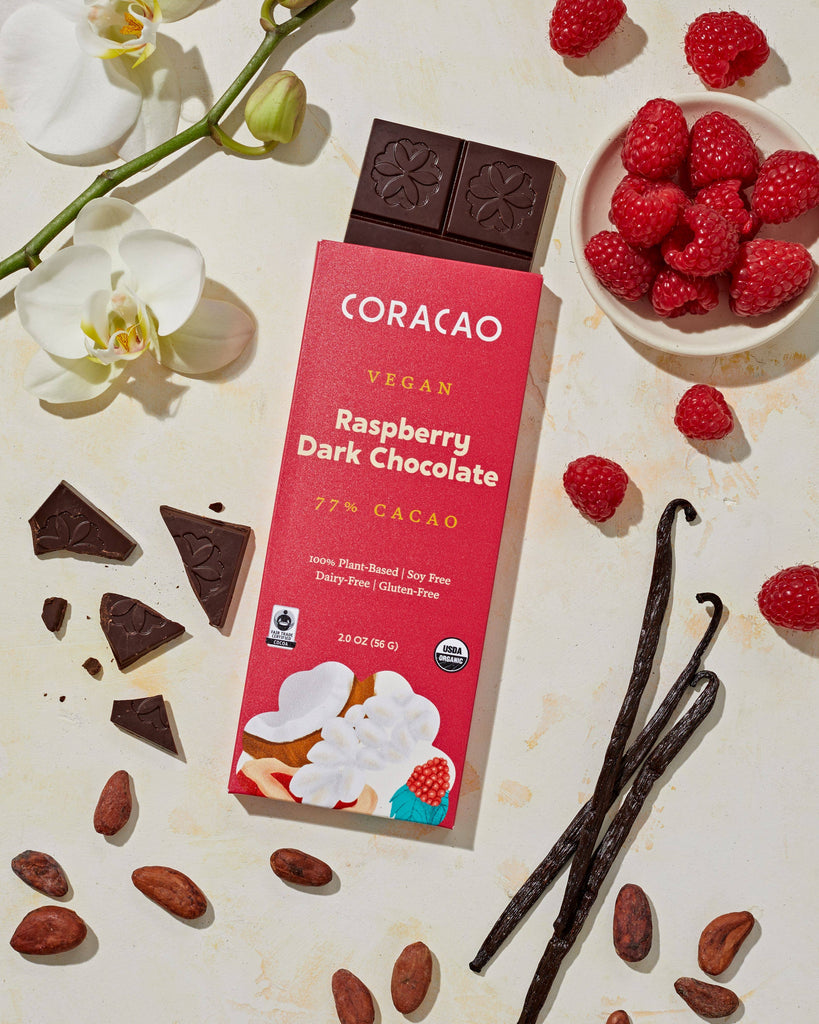 A wrapped 77% Vegan Raspberry Dark Chocolate bar on top of an unwrapped 77% Vegan Raspberry Dark Chocolate bar, with its ingredients sitting on a beige table.