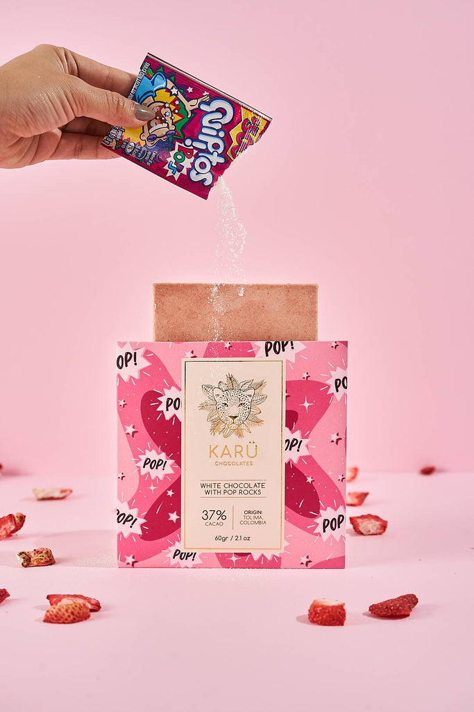 A hand pouring pop rocks on a White Chocolate with Pop Rocks 37% bar on a pink table with pieces of fruit