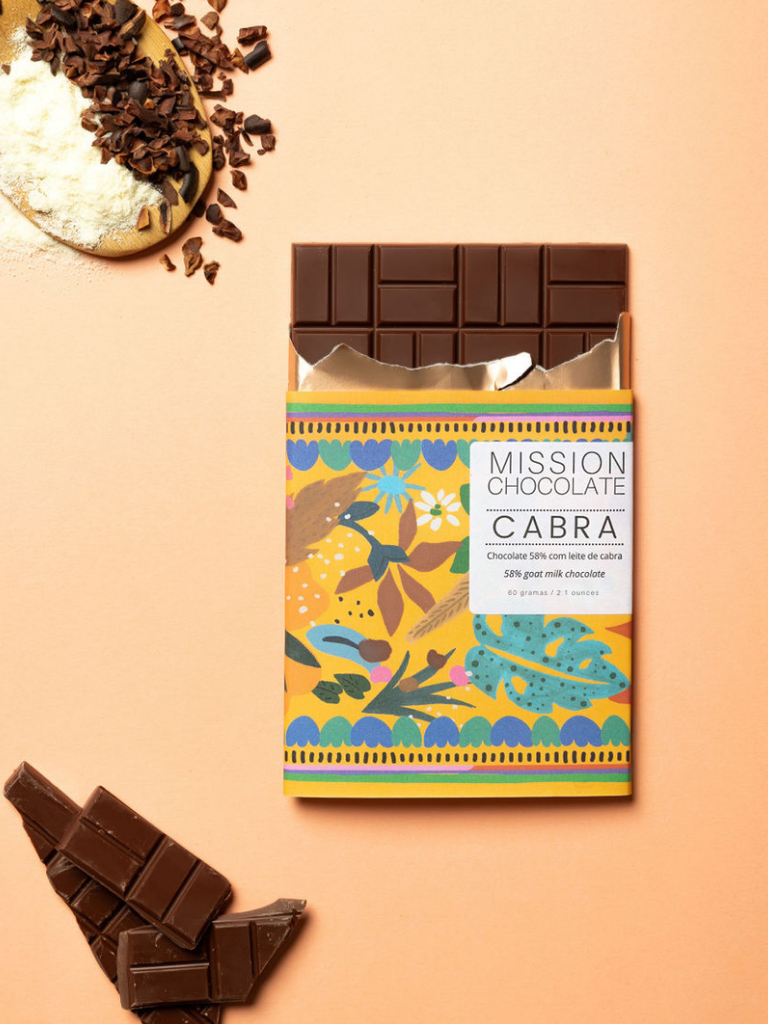 Partially opened Dark Goat Milk Chocolate 58% bar on an orange table with pieces of chocolate and ingredients on a wooden spoon