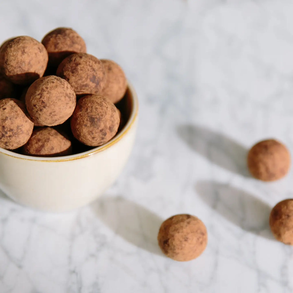 Bowl of Cocoa Dusted Marzipan Balls ona. marble table with three Cocoa Dusted Marzipan Balls on the table
