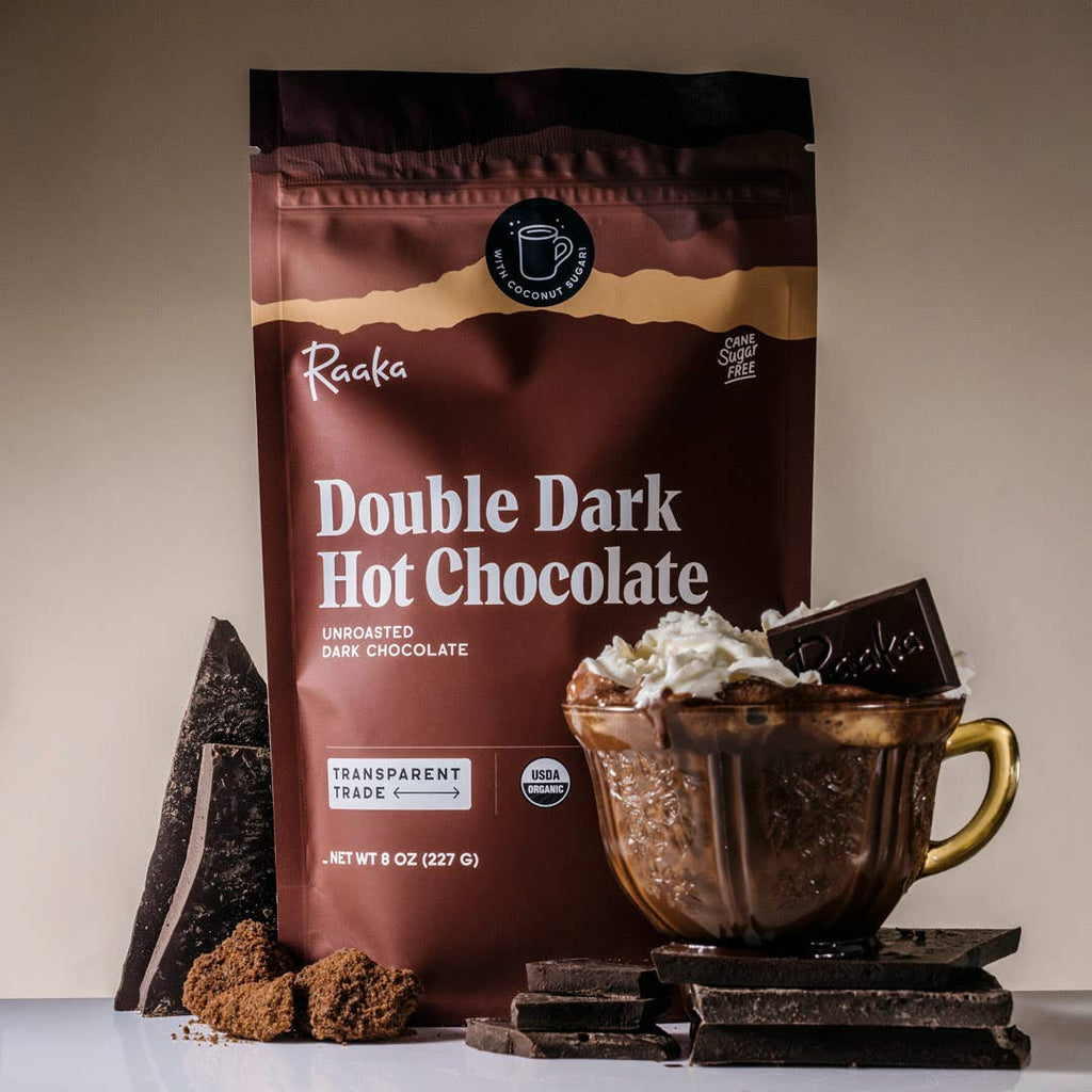A bag of Double Dark Hot Chocolate behind a mug of Double Dark Hot Chocolate topped with a piece of chocolate and whipped cream