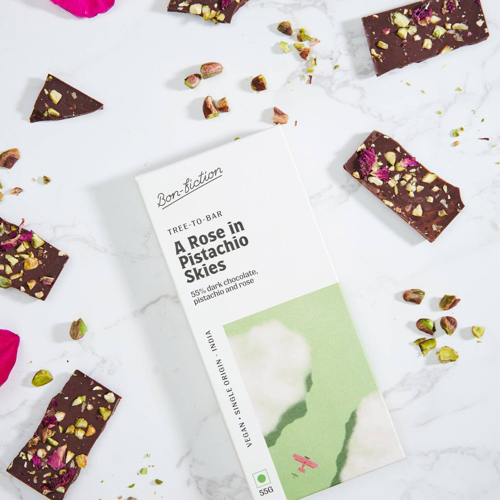 The front of a wrapped "A Rose in Pistachio Skies" chocolate bar, laying against a marble background, with pieces of the unwrapped bar scattered throughout the picture.