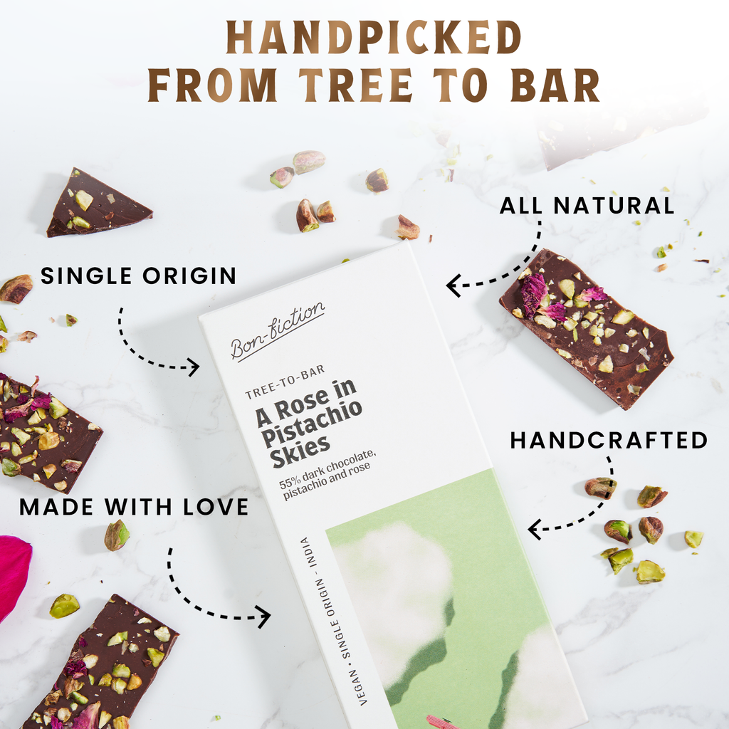 The front of a wrapped "A Rose in Pistachio Skies" chocolate bar, laying against a marble background, with pieces of the unwrapped bar scattered throughout the picture. There are "Single Origin", "Made With Love", "All Natural", and "Handcrafted" signs with arrows pointing to different parts of the unwrapped bar. 