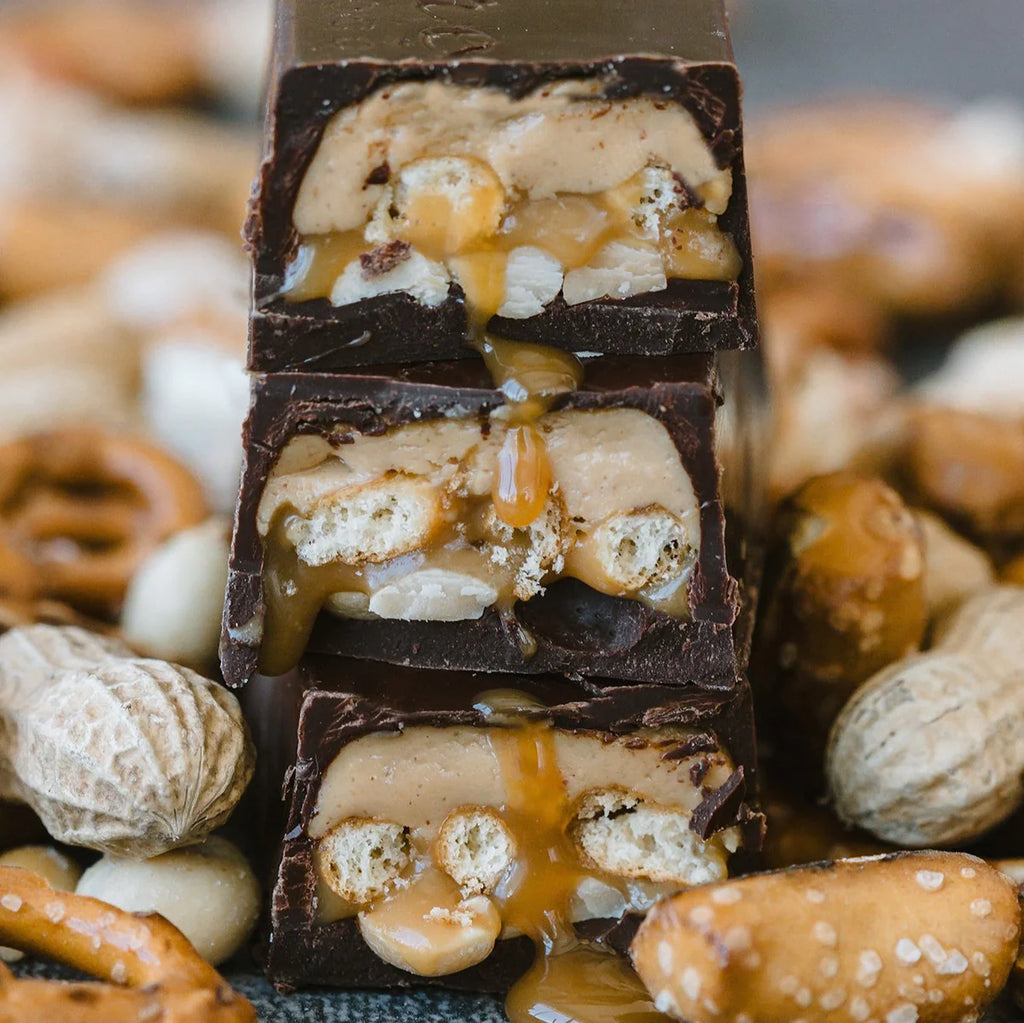 Unwrapped, stacked 5 O’Clock candy bars on a bed of peanuts, gluten-free pretzels, and other various ingredients