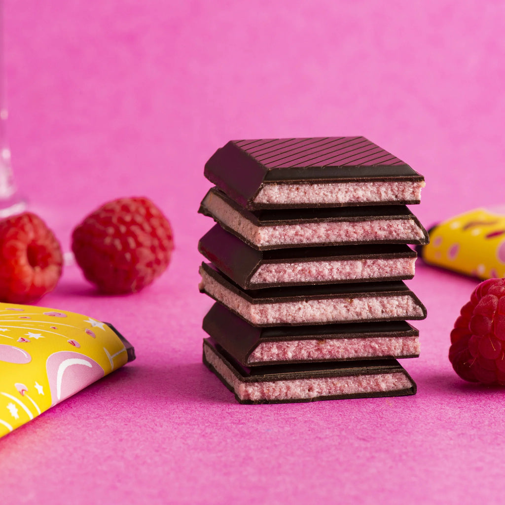 Stacked, unwrapped Sip Sip Hoorary Bars on a pink table with blurred wrapping and raspberries